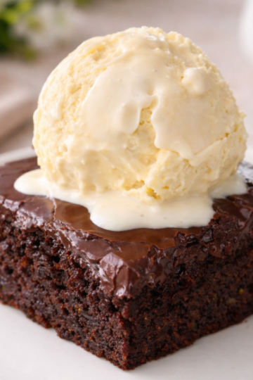Square of double chocolate zucchini cake with glossy fudge frosting and a scoop of vanilla ice cream on top, showing the moist cake–brownie hybrid texture.