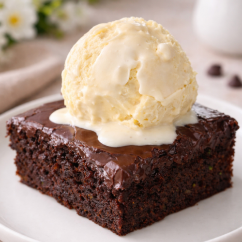 Square of double chocolate zucchini cake with glossy fudge frosting and a scoop of vanilla ice cream on top, showing the moist cake–brownie hybrid texture.