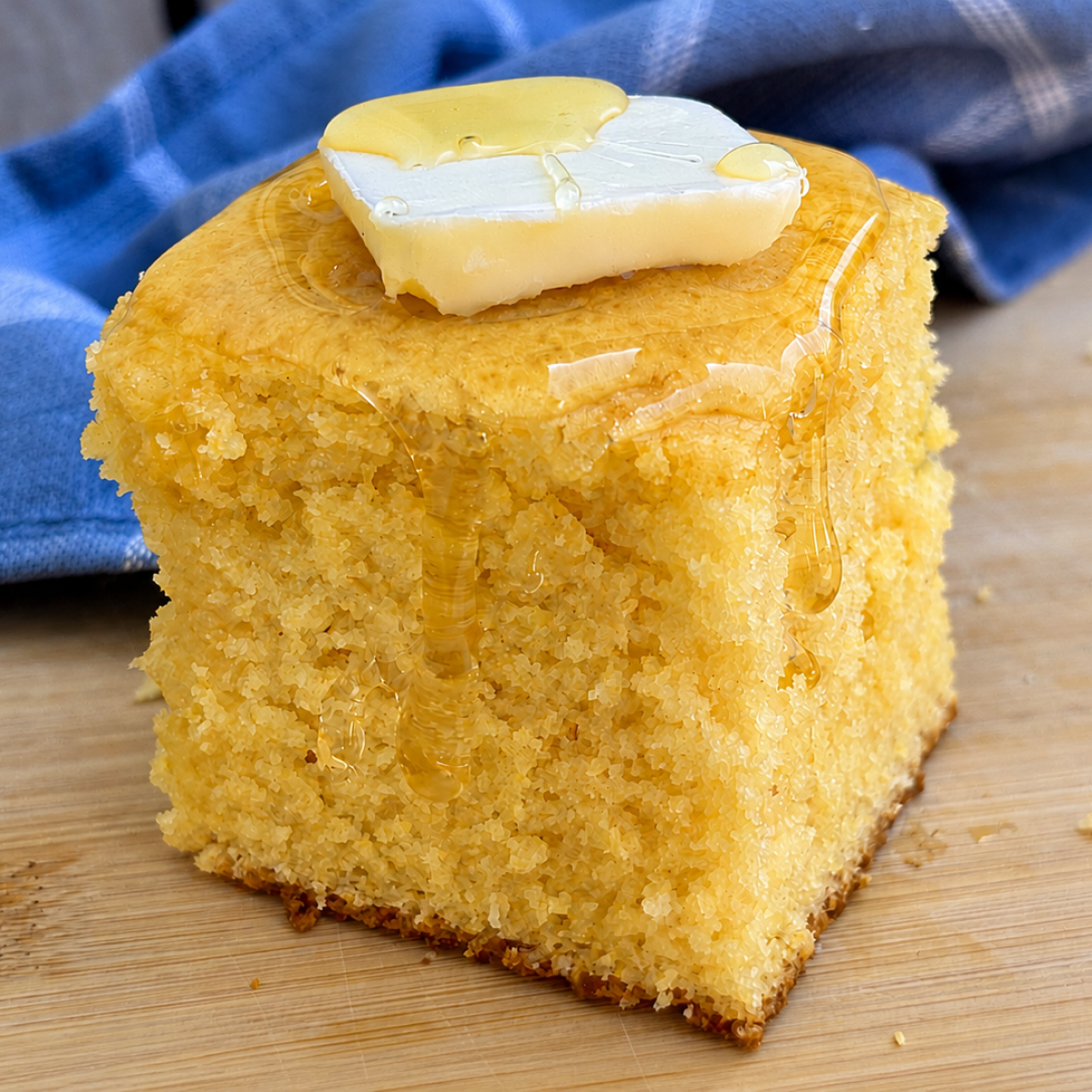Thick golden square of honey cornbread topped with a melting pat of butter and dripping honey, on a wooden cutting board with a blue linen napkin.