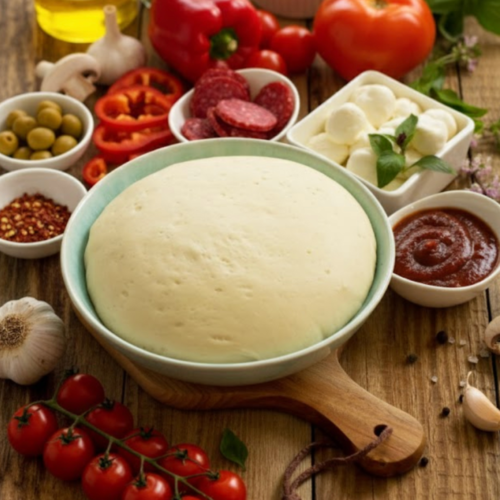 A large, fluffy ball of quick rising pizza dough in a bowl, risen to double its size surrounded by pizza toppings.