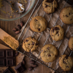 A wire cooling rack with eight gooey, golden baked brown butter chocolate chunk cookies on a rustic wooden counter.