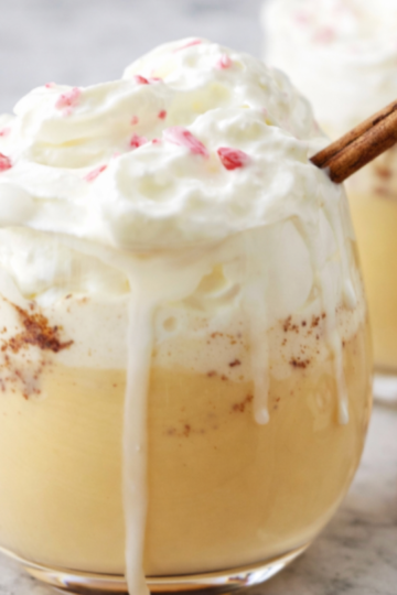 Homemade non-alcoholic eggnog topped with whipped cream and a cinnamon stick, served in a glass on a white marble countertop.