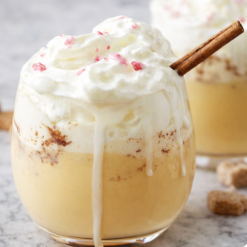 Homemade non-alcoholic eggnog topped with whipped cream and a cinnamon stick, served in a glass on a white marble countertop.