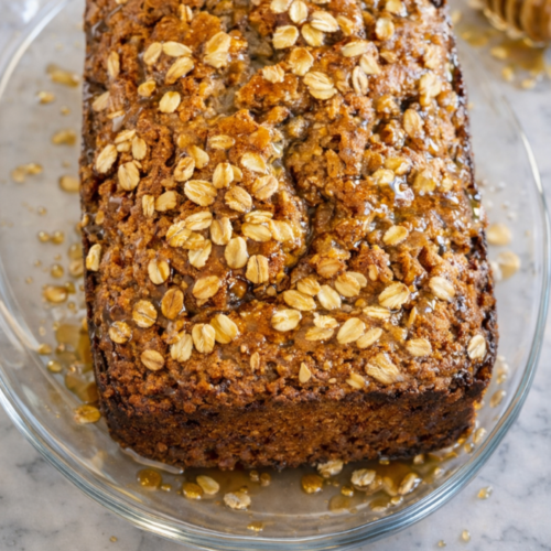 Moist chai spiced banana bread topped with rolled oats and drizzled with honey, resting on a glass plate over a white marble countertop.