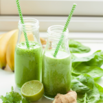 A glass of vibrant green juice with ginger and fresh mint on a light surface, surrounded by kale, banana, and lime.
