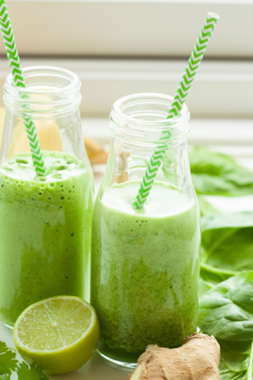 A glass of vibrant green juice with ginger and fresh mint on a light surface, surrounded by kale, banana, and lime.