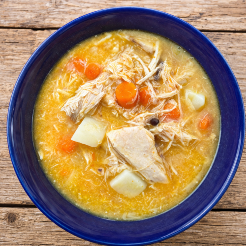 Top-down view of sourdough discard chicken soup in a blue bowl with shredded chicken, carrots, and potatoes on a rustic wooden table.