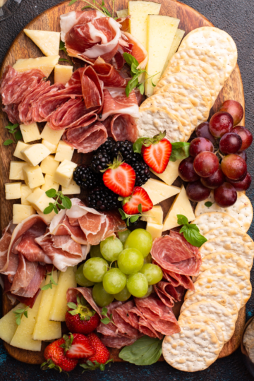 Top down view of a fully assembled, abundant charcuterie, board featuring various cheeses, cured meats, grapes, crackers, and bowls of olives and jam.