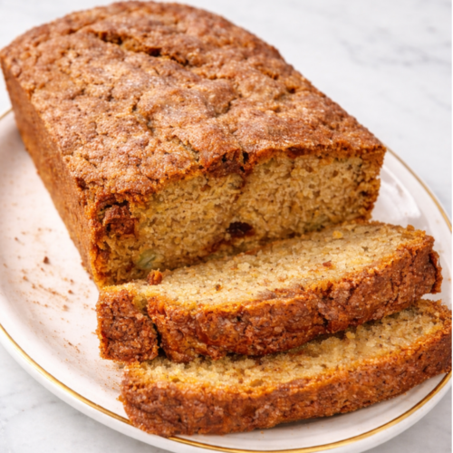 A slice of snickerdoodle banana bread with a golden cinnamon sugar crust, sitting on a wooden table with breakfast nearby.