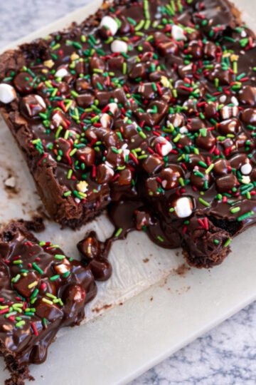 Fudgy hot chocolate brownies topped with marshmallows and cocoa frosting on a cutting board.