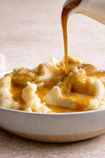 A large ceramic bowl filled with fresh hot mashed potatoes, being topped with pipping hot gravy.