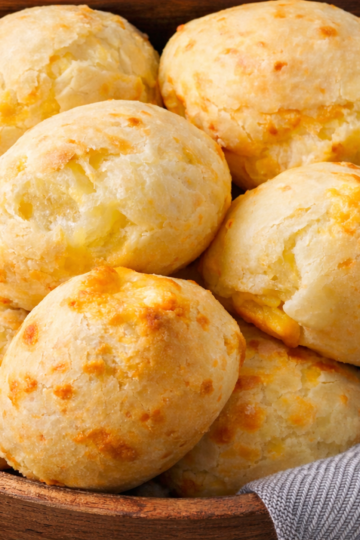 Freshly baked Brazilian cheese bread rolls arranged in a basket, showing golden cheese-speckled tops and a soft, airy texture.
