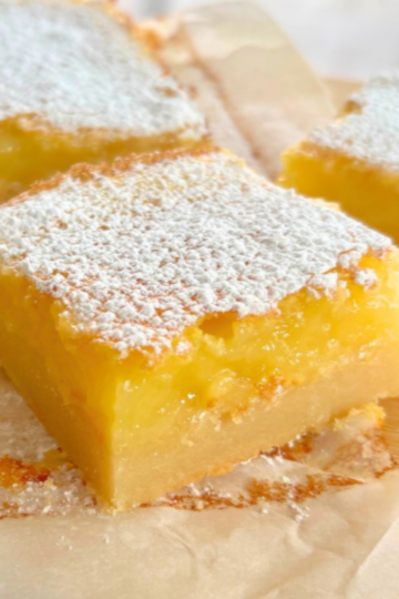 Close-up of stacked homemade lemon bars on a cutting board, dusted heavily with powdered sugar and showing thick lemon filling over a buttery shortbread crust.