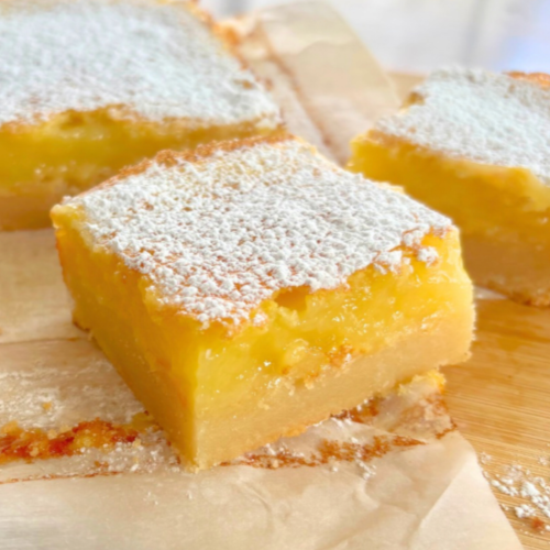 Close-up of stacked homemade lemon bars on a cutting board, dusted heavily with powdered sugar and showing thick lemon filling over a buttery shortbread crust.