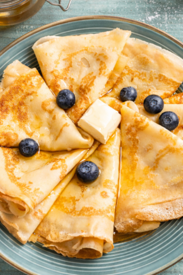 Golden sourdough discard crêpes folded into triangles and arranged on a plate with butter, honey, and fresh blueberries on a rustic wooden table.