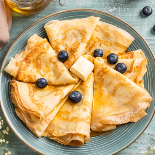Golden sourdough discard crêpes folded into triangles and arranged on a plate with butter, honey, and fresh blueberries on a rustic wooden table.