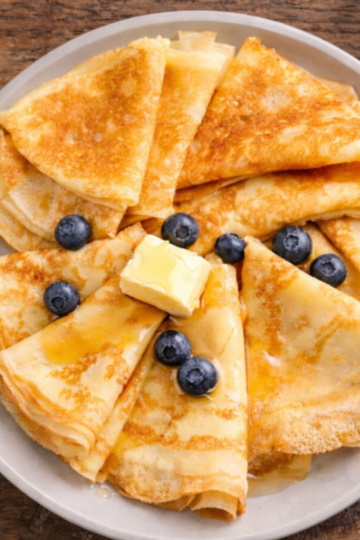 Golden sourdough discard crêpes folded into triangles and arranged on a plate with butter, honey, and fresh blueberries on a rustic wooden table.