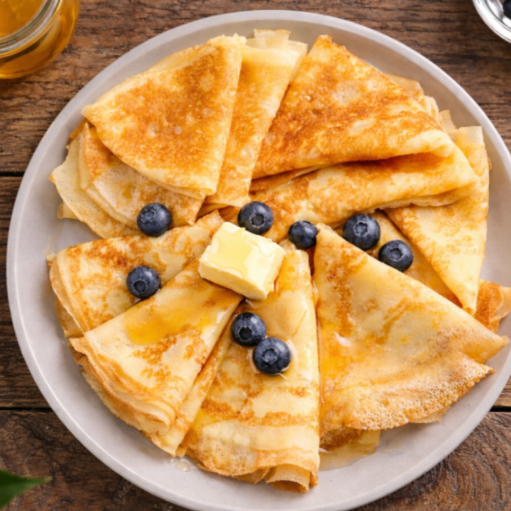 Golden sourdough discard crêpes folded into triangles and arranged on a plate with butter, honey, and fresh blueberries on a rustic wooden table.