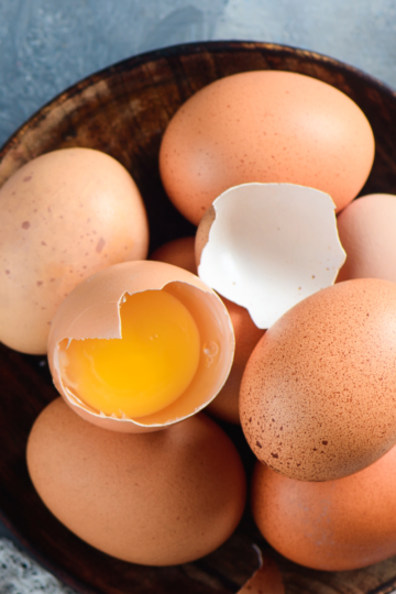 A nice wooden bowl filled with brown chicken eggs, one being broken half open to show the raw yolk.