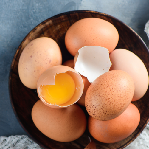A nice wooden bowl filled with brown chicken eggs, one being broken half open to show the raw yolk.