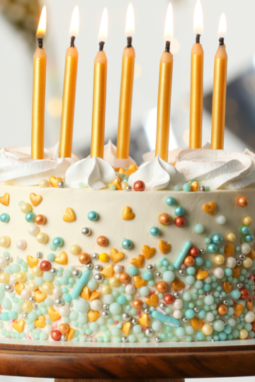 A simple birthday cake decorated with sprinkles and topped with lit candles resting on a wooden cake stand.