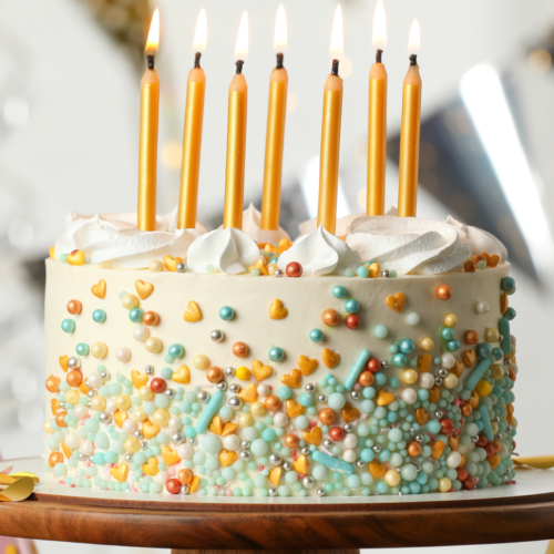 A simple birthday cake decorated with sprinkles and topped with lit candles resting on a wooden cake stand.