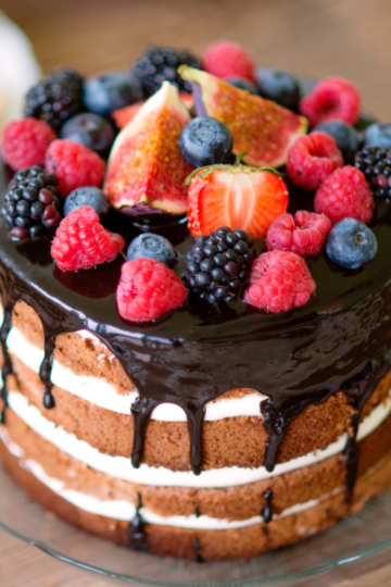 A beautiful cake with chocolate frosting dripping down the sides topped with fresh colorful fruit, served on a glass plate.