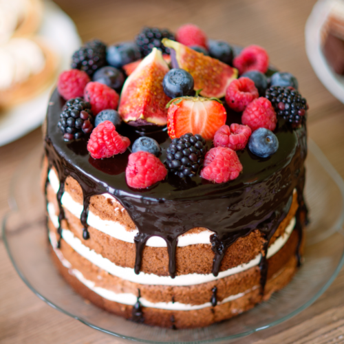 A beautiful cake with chocolate frosting dripping down the sides topped with fresh colorful fruit, served on a glass plate.