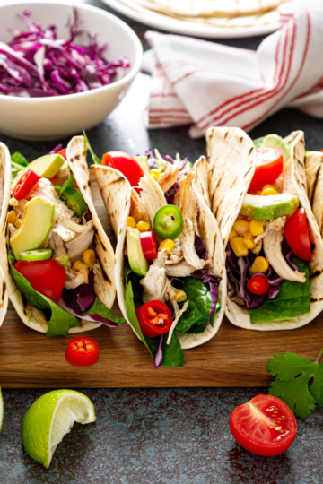 A nice wooden cutting board filled with flour tortilla tacos with colorful filling spilling out of them.