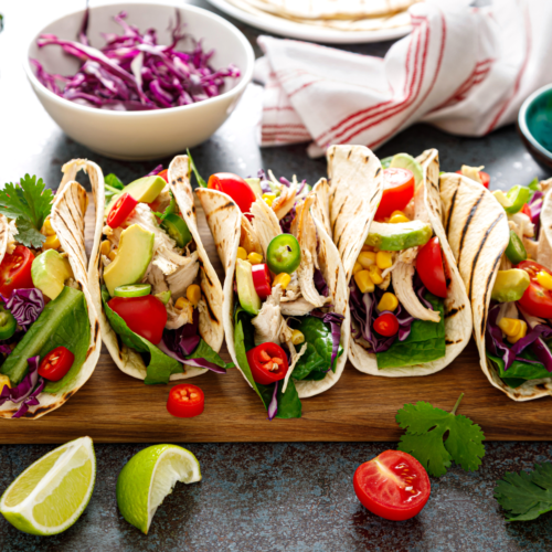 A nice wooden cutting board filled with flour tortilla tacos with colorful filling spilling out of them.