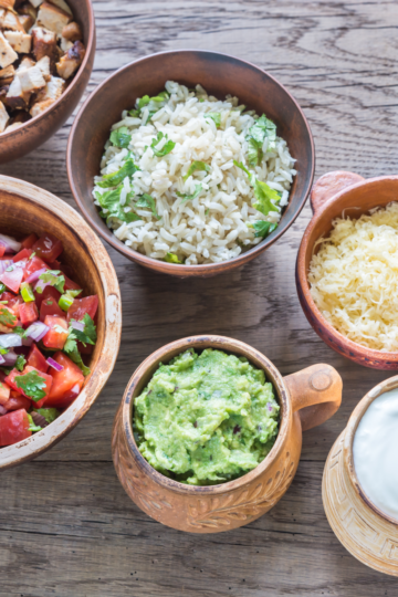 Overhead view of taco bar ingredients in wooden bowls including diced chicken, cilantro rice, pico de gallo, shredded cheese, black beans, guacamole, sour cream, and peppers with fresh limes.