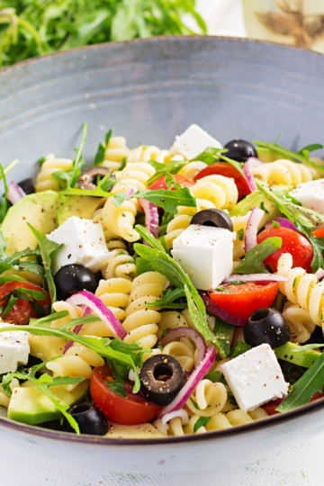 A ceramic bowl filled with colorful pasta salad including arugula, olives, feta and spiral pasta.