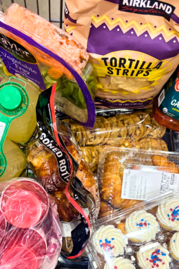 Costco shopping cart loaded with graduation party food including Kirkland tortilla strips, rotisserie chicken, croissants, vegetable tray, lemonade, Jack's Cantina salsa, chocolate chip cookies, and red, white, and blue cupcakes.