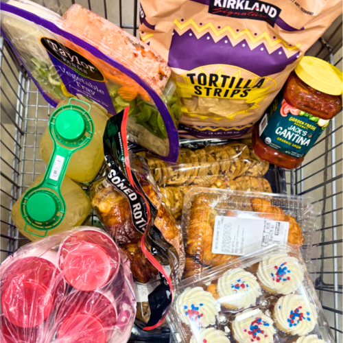 Costco shopping cart loaded with graduation party food including Kirkland tortilla strips, rotisserie chicken, croissants, vegetable tray, lemonade, Jack's Cantina salsa, chocolate chip cookies, and red, white, and blue cupcakes.
