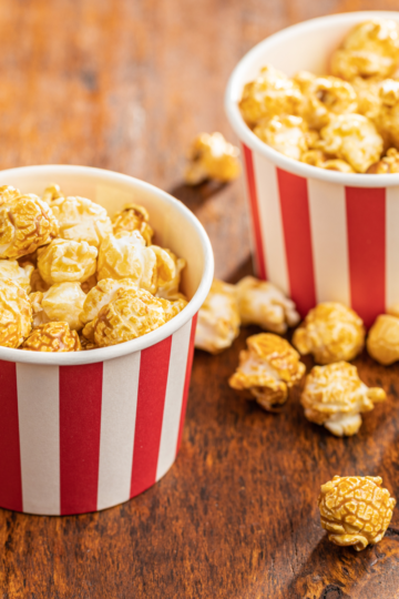 Two red and white striped popcorn cups filled with golden caramel popcorn on a rustic wooden table, with extra popcorn pieces scattered around.