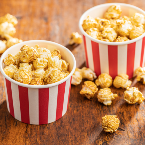 Two red and white striped popcorn cups filled with golden caramel popcorn on a rustic wooden table, with extra popcorn pieces scattered around.
