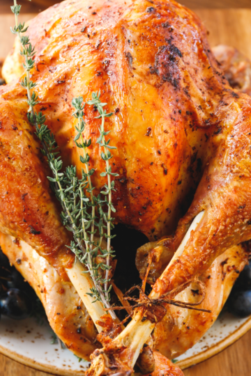 Golden roasted whole Thanksgiving turkey on a wooden cutting board, garnished with fresh thyme and surrounded by black grapes and persimmons.