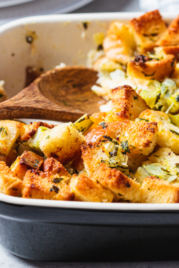 Golden homemade stuffing with toasted bread cubes, celery, and fresh herbs in a baking dish with a wooden serving spoon.