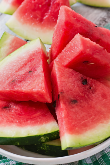 Fresh watermelon wedges with the rind on, stacked on a white plate over a green gingham napkin.