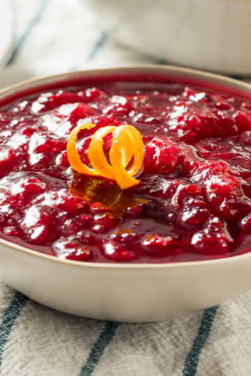 Bowl of homemade cranberry sauce garnished with an orange twist on a striped napkin.