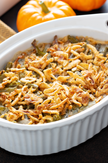 Green bean casserole topped with crispy fried onions in a white oval baking dish with pumpkins in the background.