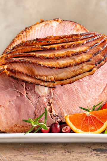 Glazed spiral-cut bone-in ham on a white platter garnished with orange slices, cranberries, and rosemary