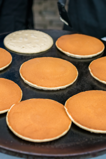 Eight golden pancakes cooking on a large round griddle with a pitcher of batter beside it.