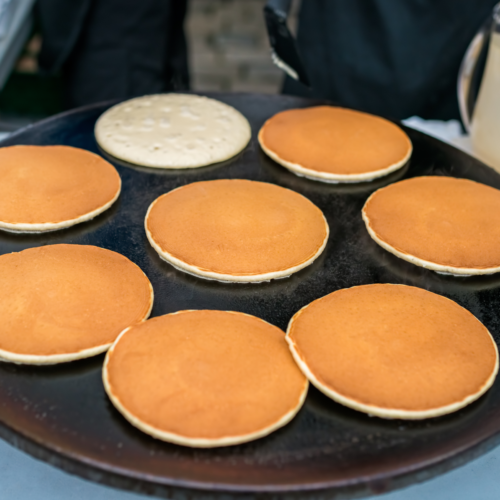Eight golden pancakes cooking on a large round griddle with a pitcher of batter beside it.