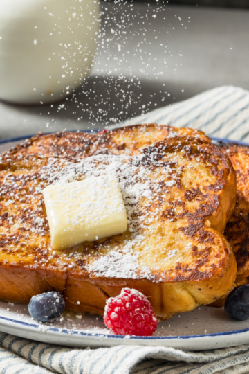 Three thick slices of brioche French toast on a white plate with butter, powdered sugar, and fresh berries
