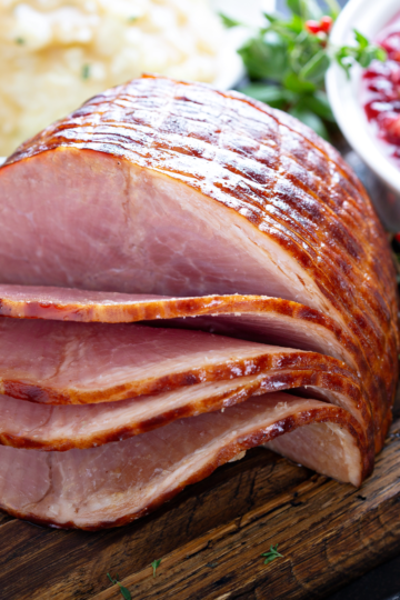 Spiral-sliced glazed ham on a wooden board with mashed potatoes and cranberry sauce behind it.